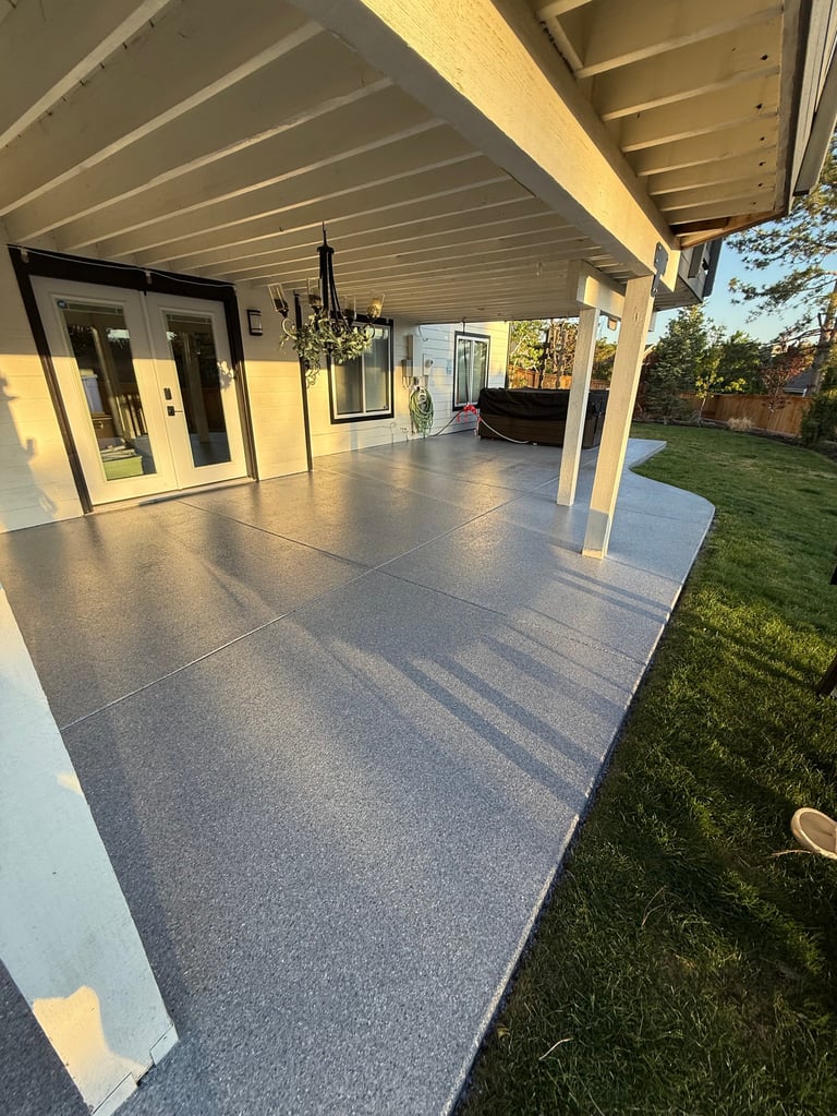 Modern covered patio with gray concrete flooring, white columns, and white ceiling attached to a contemporary house with green lawn visible on the right